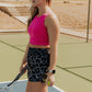 blonde haired women standing in the middle of a tennis court. she is holding a tennis racket in one hand and a tennis ball in the other. She is wearing a bright pink long lined tank top and a pair of printed running shorts in a panther printing. These are high waisted and have a built in panty liner.