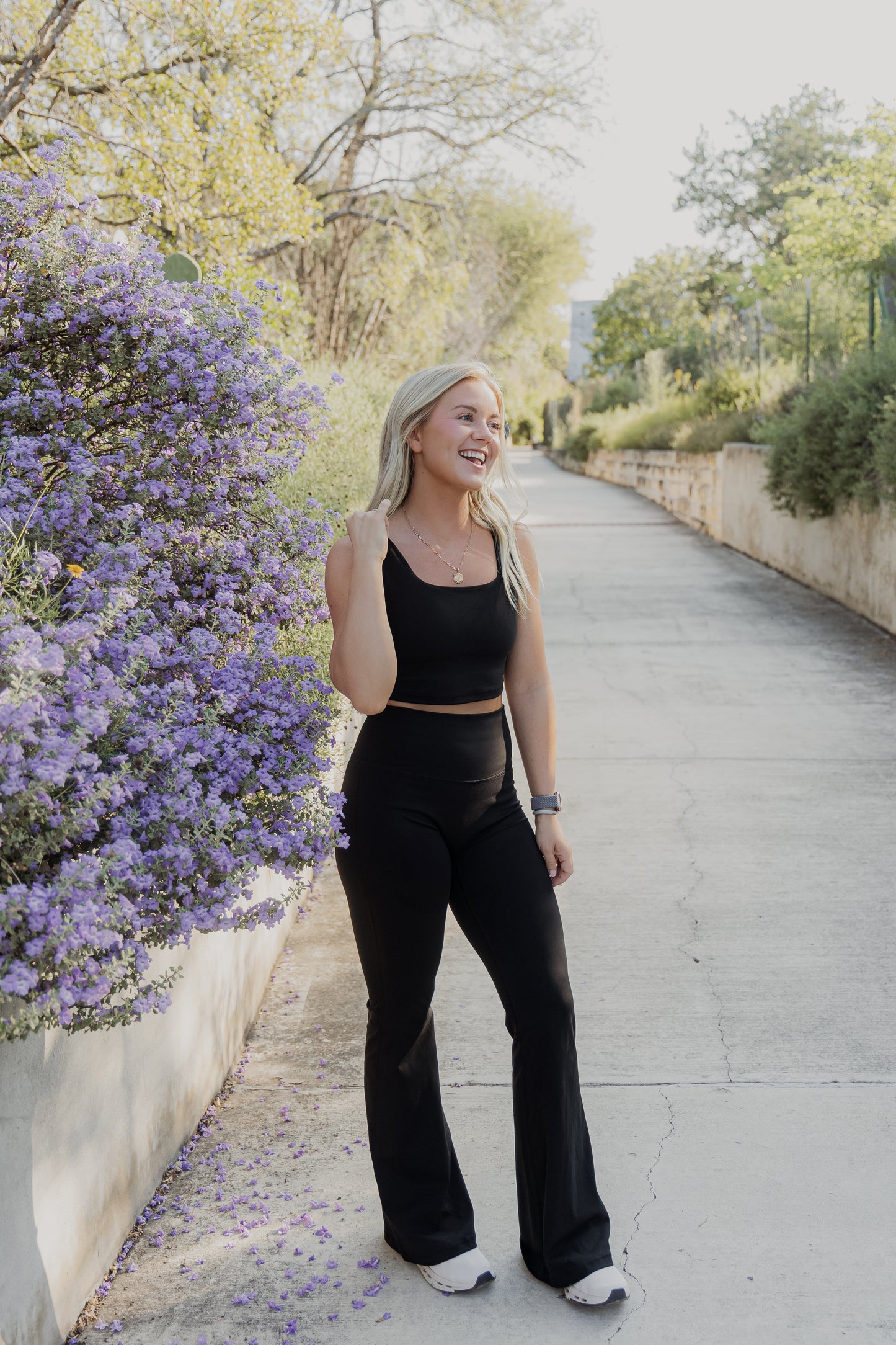 White women with blonde hair wearing a black tank top and flare leggings. She is messing with her tank top while smiling at someone off the photo.