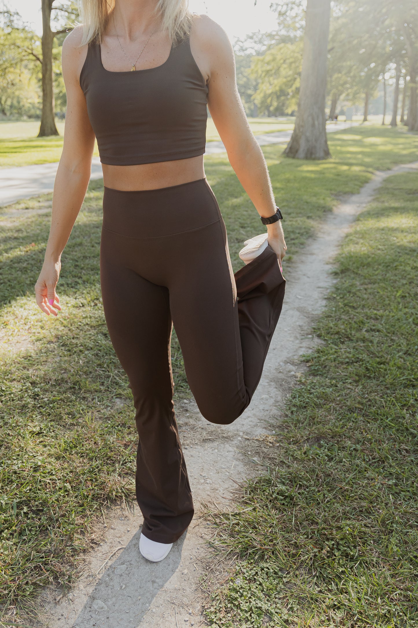 White women with blonde hair wearing a brown tank top and flare leggings. She is stretching her leg. Her face is not pictured.