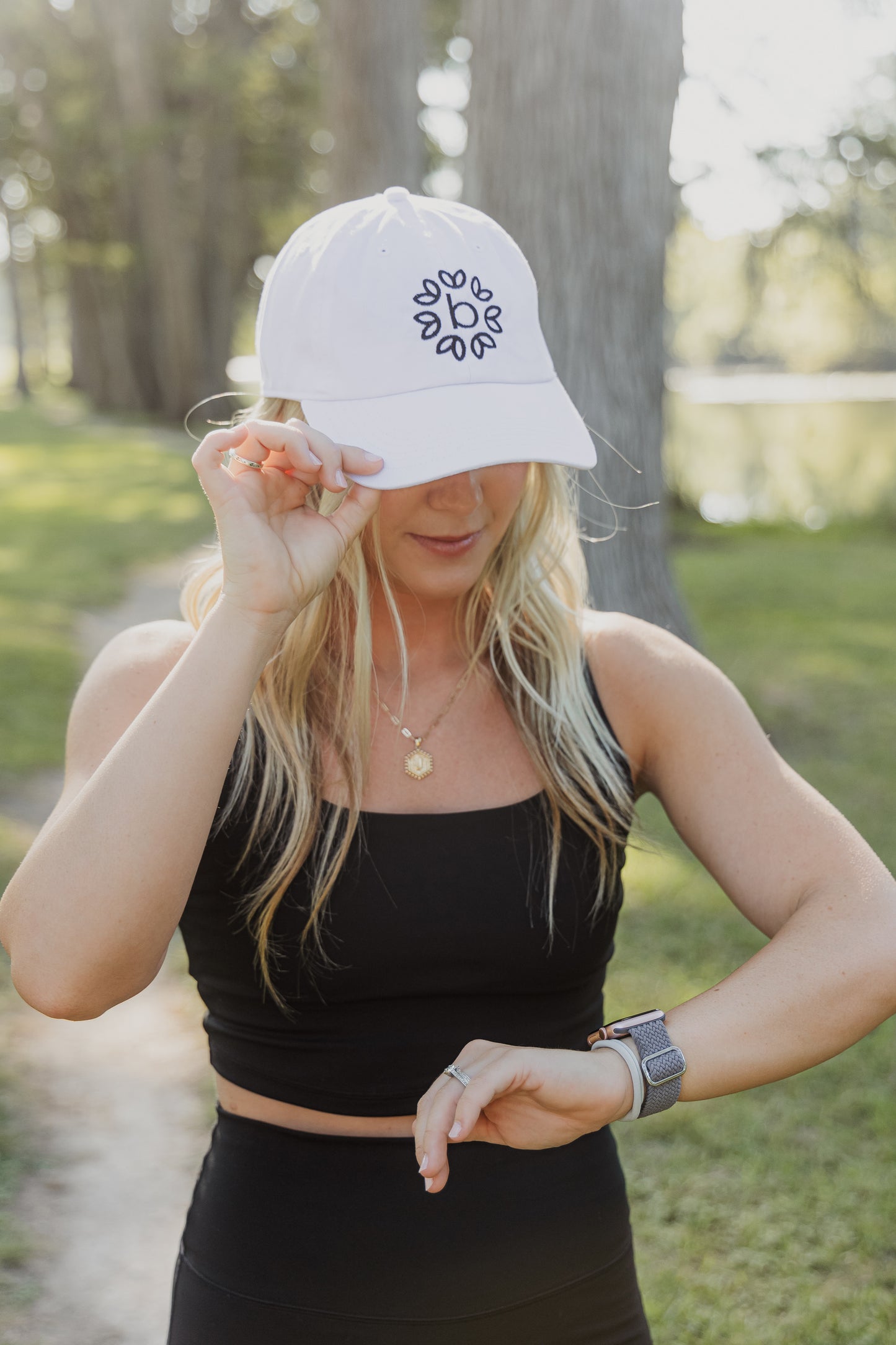 White women with blonde hair wearing a white baseball cap with a logo on the front of it. She is standing in nature and looking at her watch while holding the hat with one hand as well.