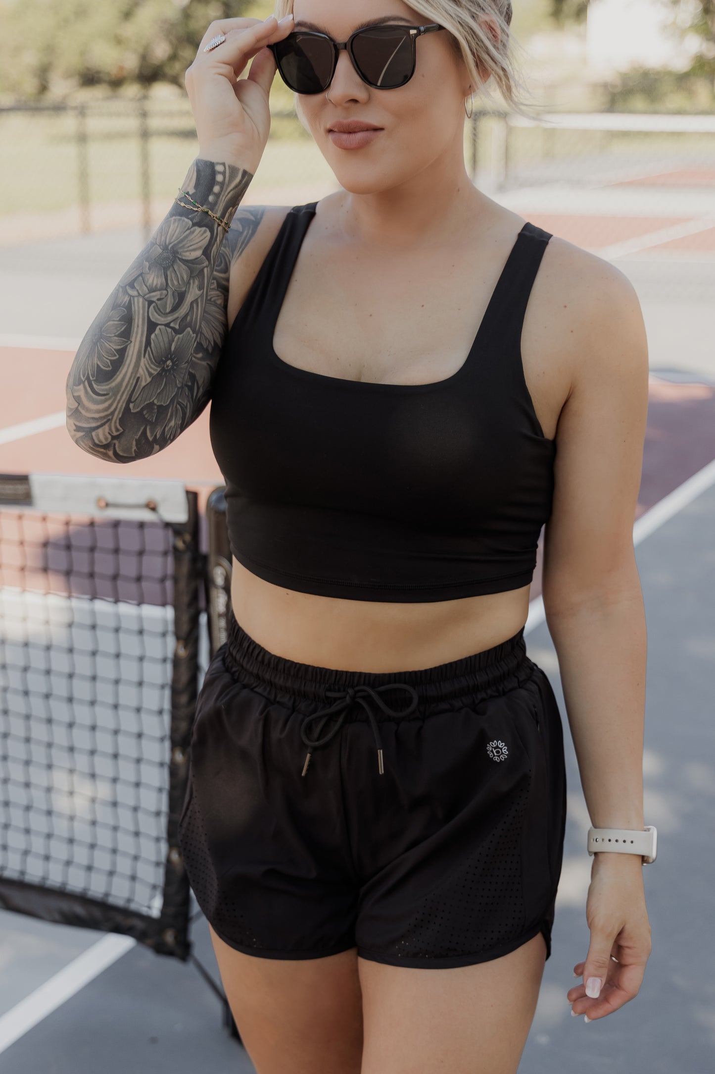 White women with blonde hair that is put up in a messing bun, she is standing on a pickle ball court wearing a black tank top sports bra and black running shorts. she is messing with her sunglasses that are on her face.
