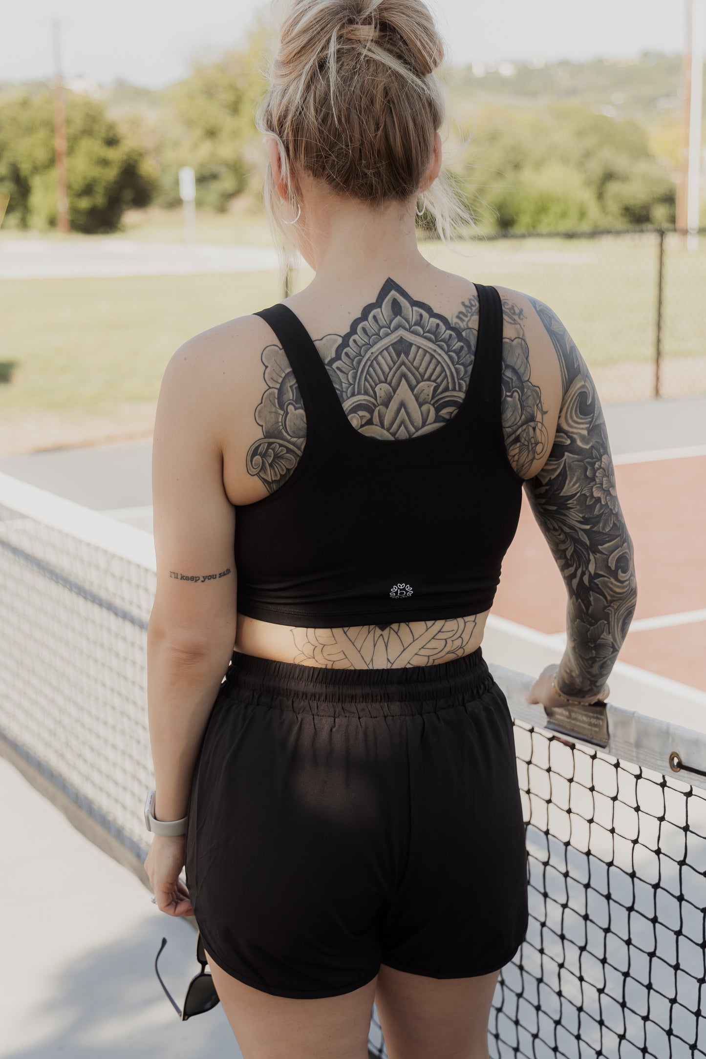 White women with blonde hair that is put up in a messy bun. She is facing away from the camera showing off her back side. She is wearing a black tank top sports bra and black running shorts. She is holding a pair of sunglasses in her left hand and has her right hand resting on the pickle ball net while standing on a pickle ball court.