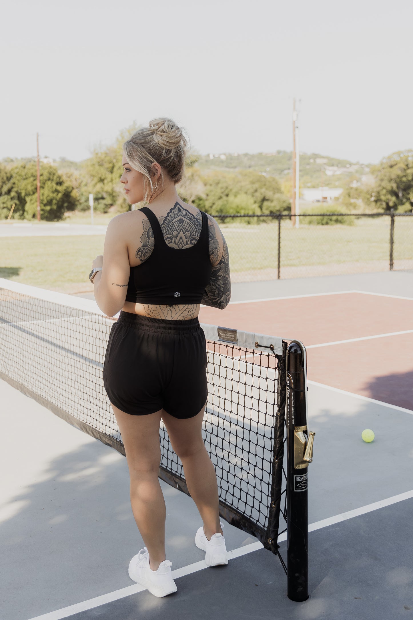 White women with blonde hair that is put up in a messy bun. She is facing away from the camera showing off her back side. She is wearing a black tank top sports bra and black running shorts. She is looking over her shoulder but not at the camera