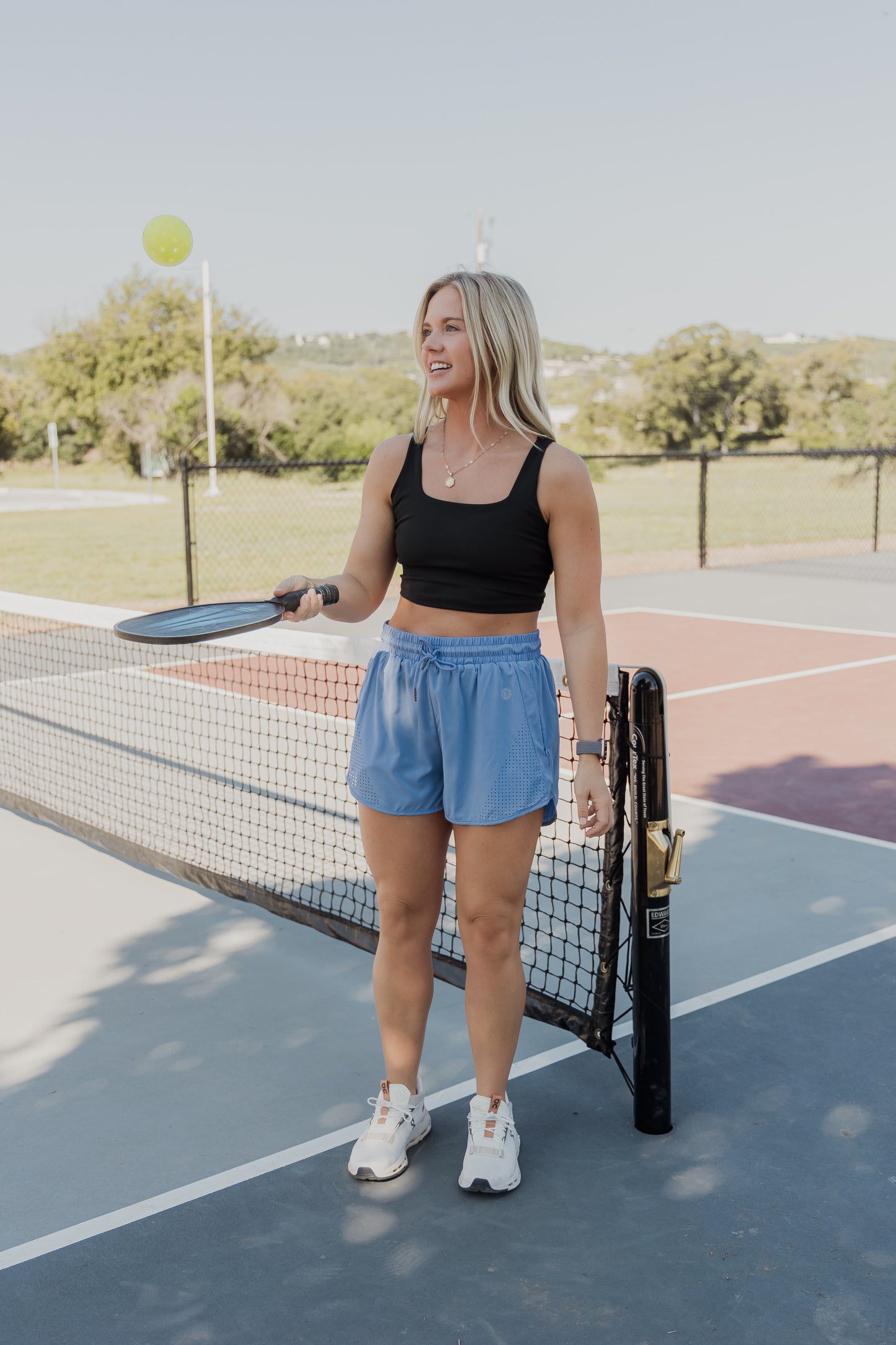 White women with blonde hair wearing a black tank top sports bra and blue running shorts, and white tennis shoes. She is holding a pickle ball paddle and trying to keep the ball up. She is standing on a pickle ball court with the net in the background.