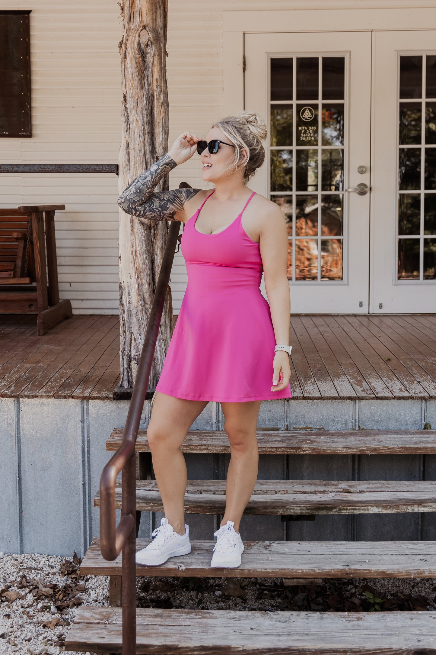 White women with blonde hair wearing it in a messy bun. She is wearing a pink tennis dress and white van athletic shoes. She is adjusting her sunglasses on her face. She is not looking at the camera and is standing on a set of wood and metal stairs infront of a white building with double doors.