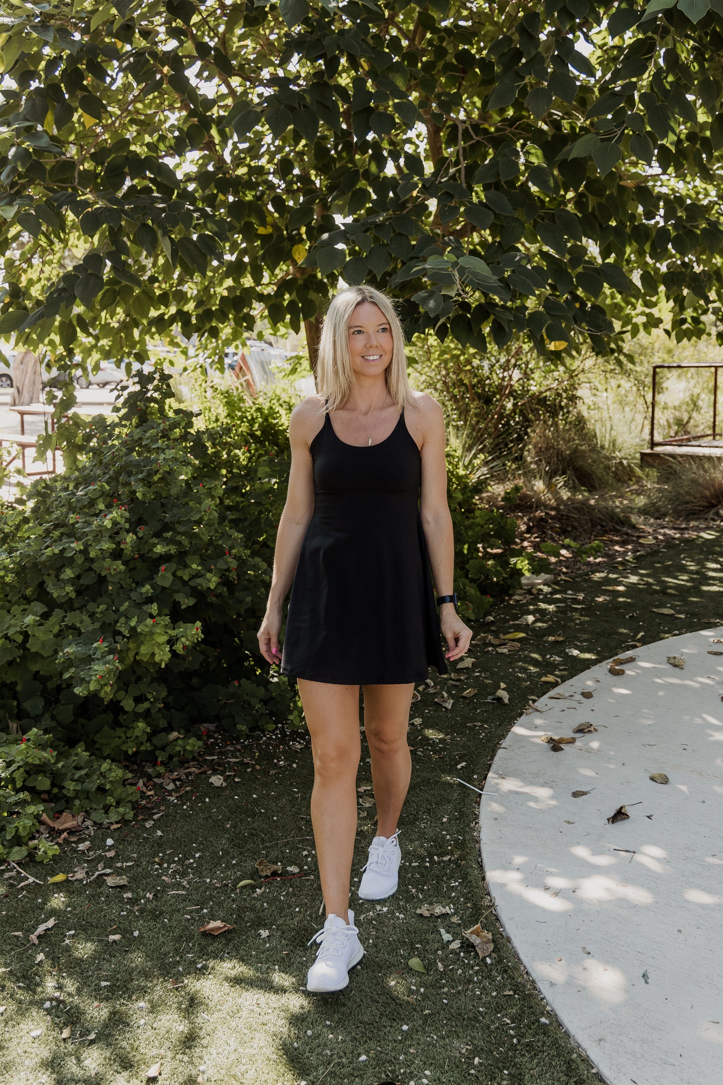 White women with blonde hair wearing a black tennis dress with white shoes. She is walking towards the camera with her arms by her side and looking off into the distance with a smile on her face.