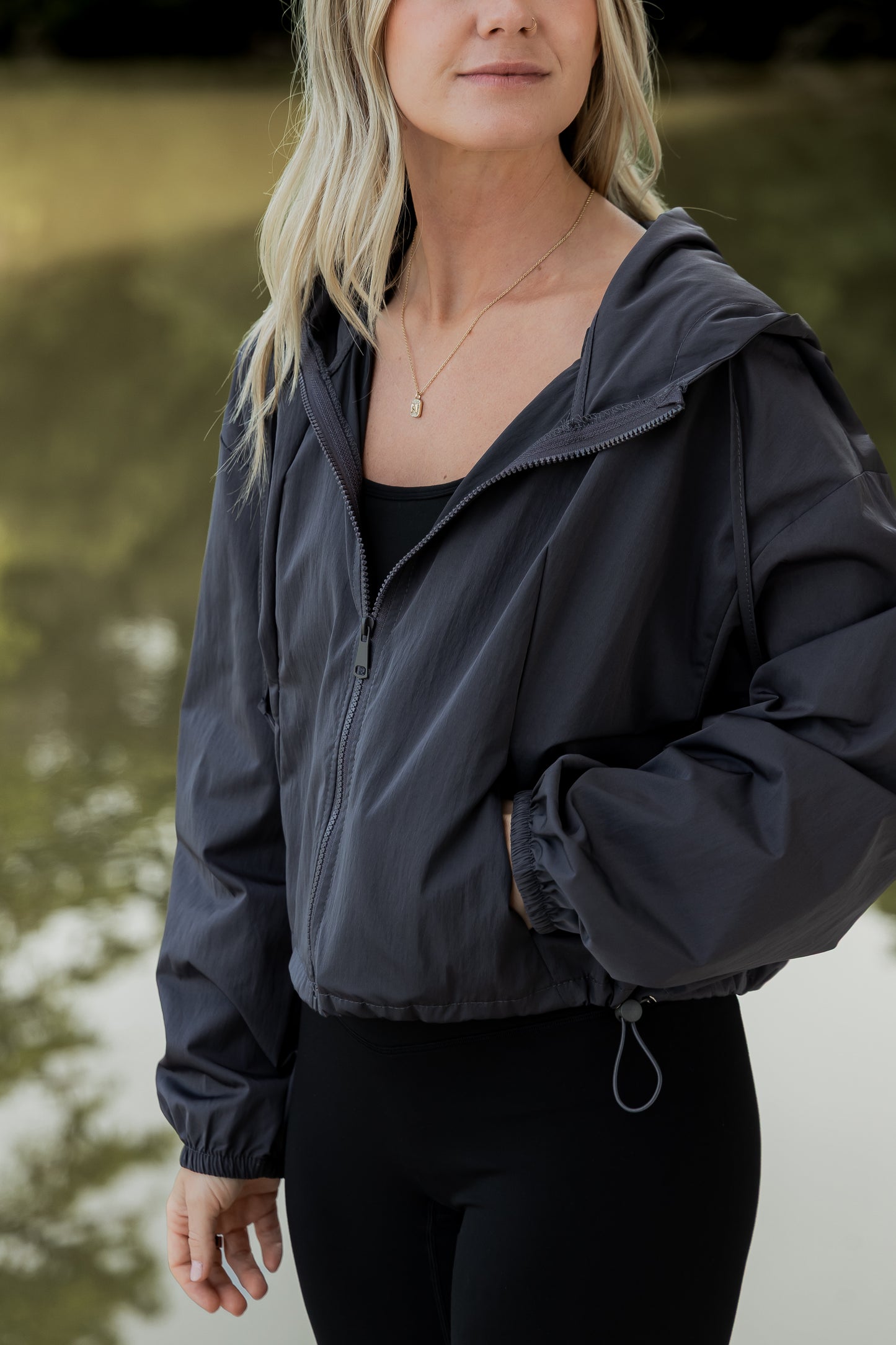 White women with blonde hair wearing a grey cropped hooded wind/rain jacket. She is facing diagonal from the camera and is not showing her whole face. Mainly its just an upclose shot of the jacket.