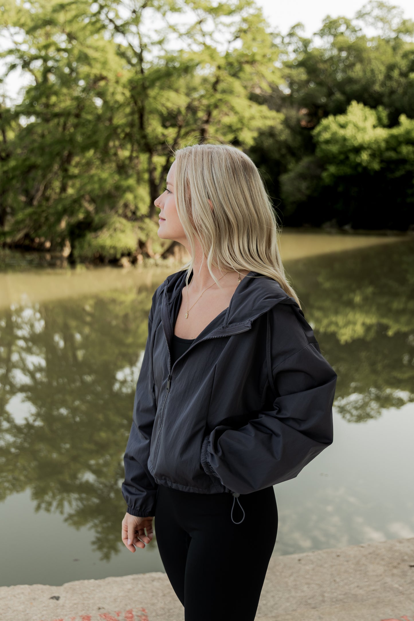 White women with blonde hair wearing a grey cropped hooded wind/rain jacket. She is faced away from the camera and is standing infront of a river at the park.