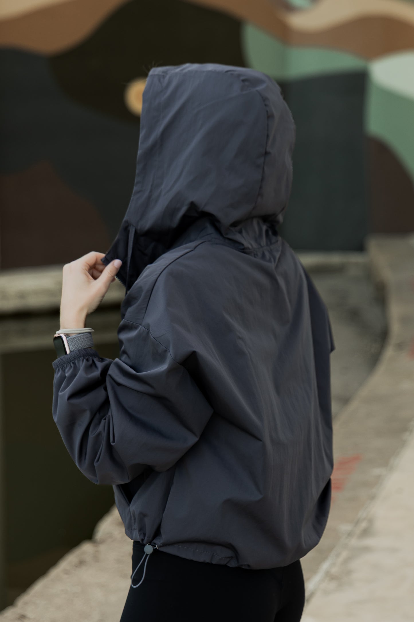 White women with blonde hair wearing a grey cropped hooded wind/rain jacket. She is facing away from the camera and is showing off the hood on the jacket. You cannot see her face at all.