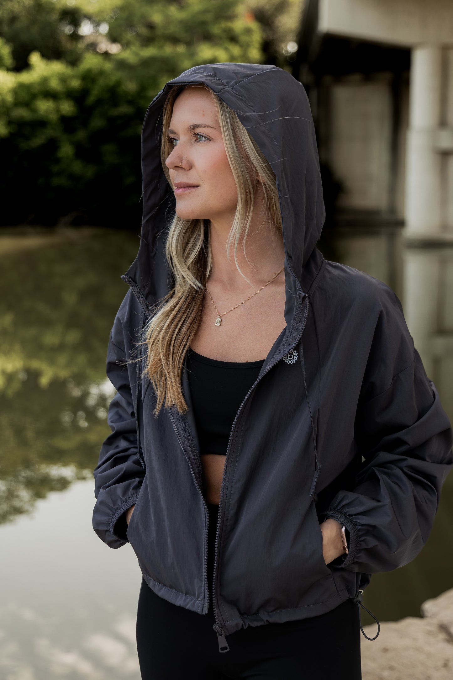 White women with blonde hair wearing a grey cropped hooded wind/rain jacket. She has the hood over her head and is shown looking off into the distance.