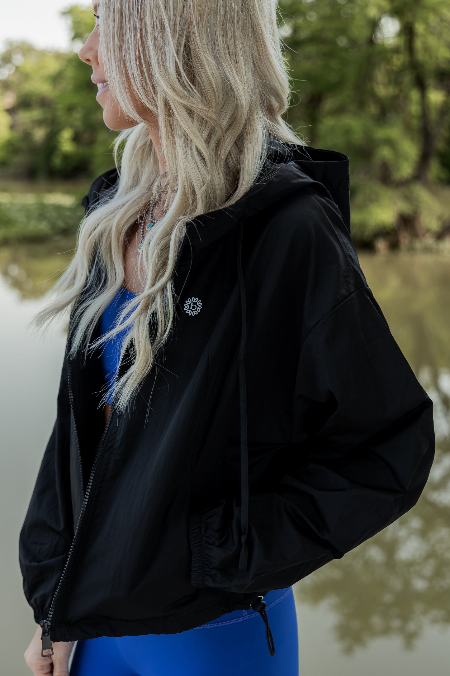 White women with blonde hair wearing a black cropped hooded wind/rain jacket. She is showing off the pocket by putting her hand in it.