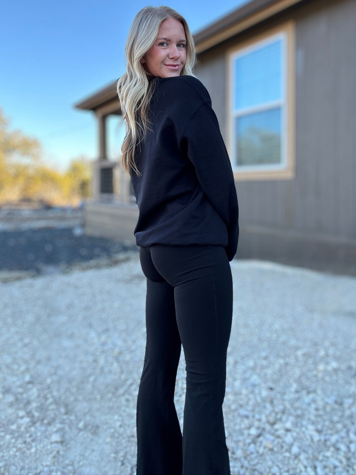 White blonde haired woman wearing a black gilden sweatshirt. She is looking back over her shoulder and showing off the sweatshirt that is paired witha pair of black flare leggings. The sweatshirt has a black logo embroidered on it with our company logo.