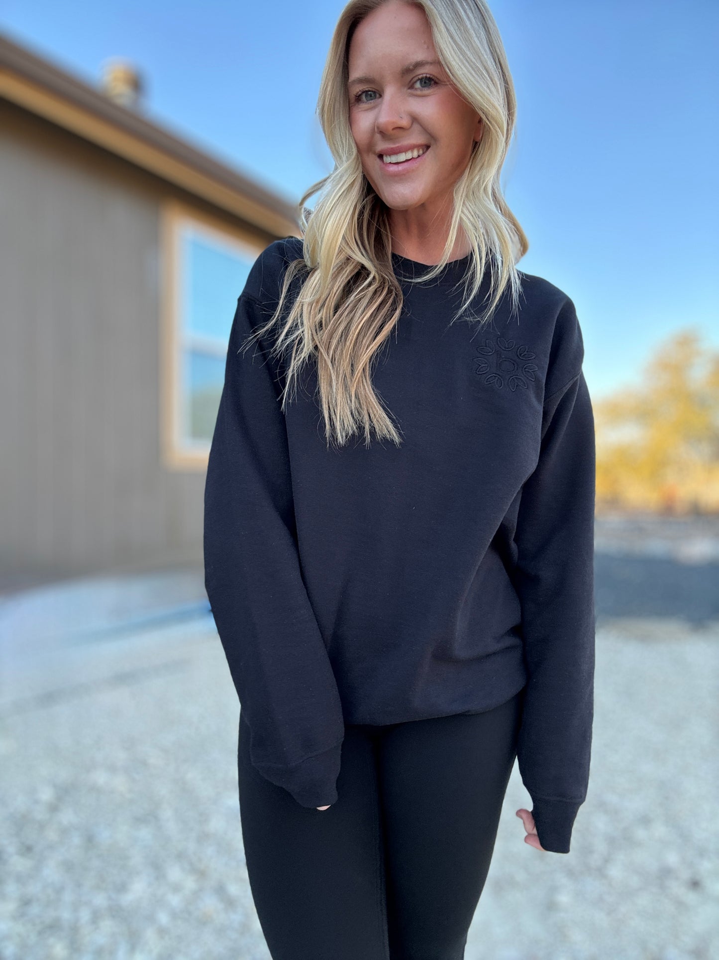 White blonde haired woman wearing a black gilden sweatshirt. She is looking at the camera and showing off the sweatshirt. The sweatshirt has a black logo embroidered on it with our company logo.