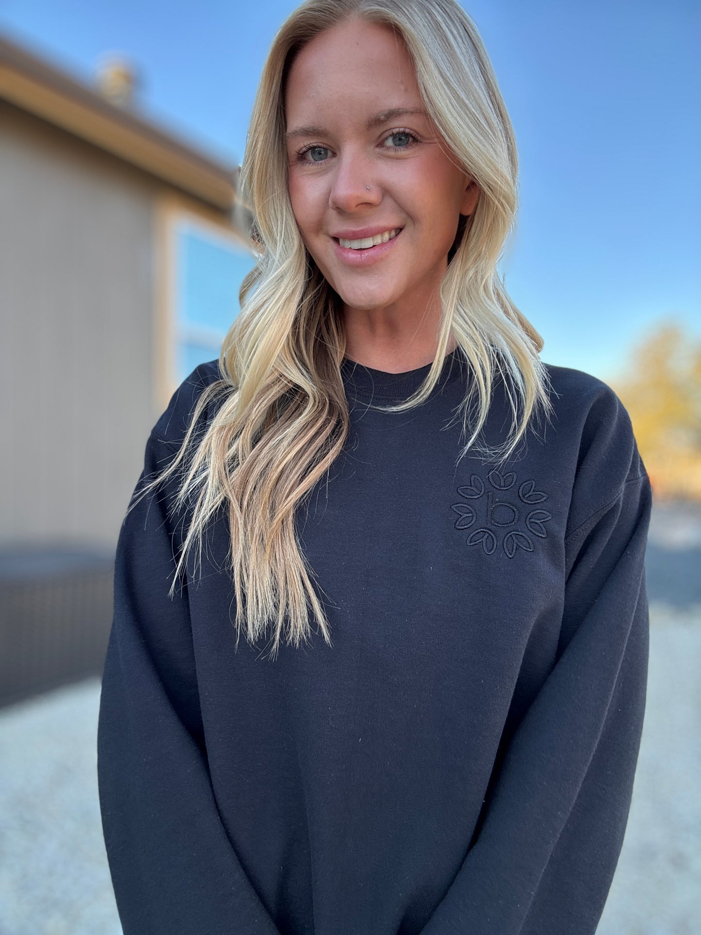 White blonde haired woman wearing a black gilden sweatshirt. She is looking at the camera and showing off the sweatshirt. The sweatshirt has a black logo embroidered on it with our company logo.