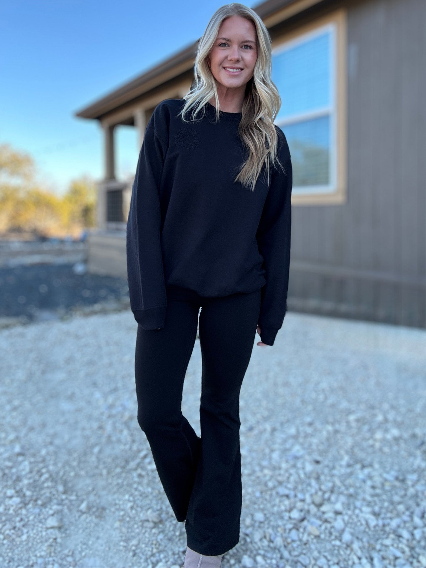 White blonde haired woman wearing a black gilden sweatshirt. She is looking at the camera and showing off the sweatshirt that is paired with a pair of black flare leggings. The sweatshirt has a black logo embroidered on it with our company logo.