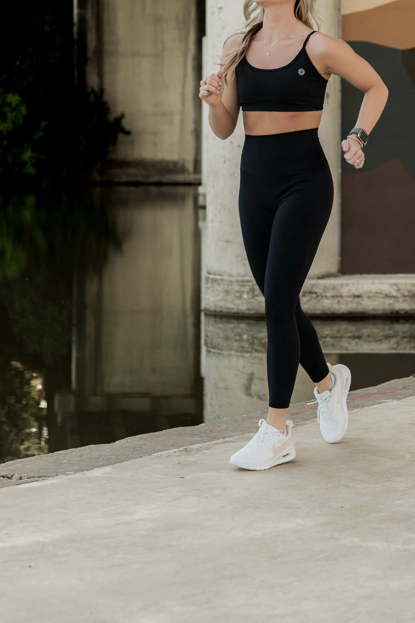 White blonde women with blonde hair running by the water at the park. she is wearing a black matching leggings and sports bra set and white tennis shoes.