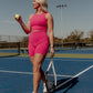 Blonde haired women walking on the middle of a tennis court holding a tennis ball and racket.  She is looking away from the camera as she is walking by.  She is wearing a matching workout set which includes a long lined tank top (with a builtin bra) and a pair of matching biker shorts in the color bright pink.