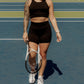 Blonde haired women walking on a tennis court with a tennis racket in her hand.  She is facing the camera and has her hair up.  She is wearing a matching tank top and biker short set in the color black.