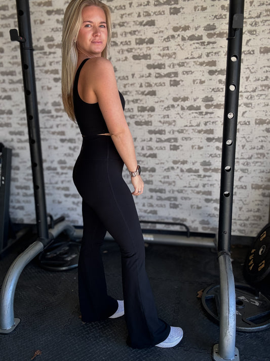 white blonde women standing in front of a white washed brick wall.  she is wearing a pair of black flare leggings and a matching long lined tank top with white sneakers.