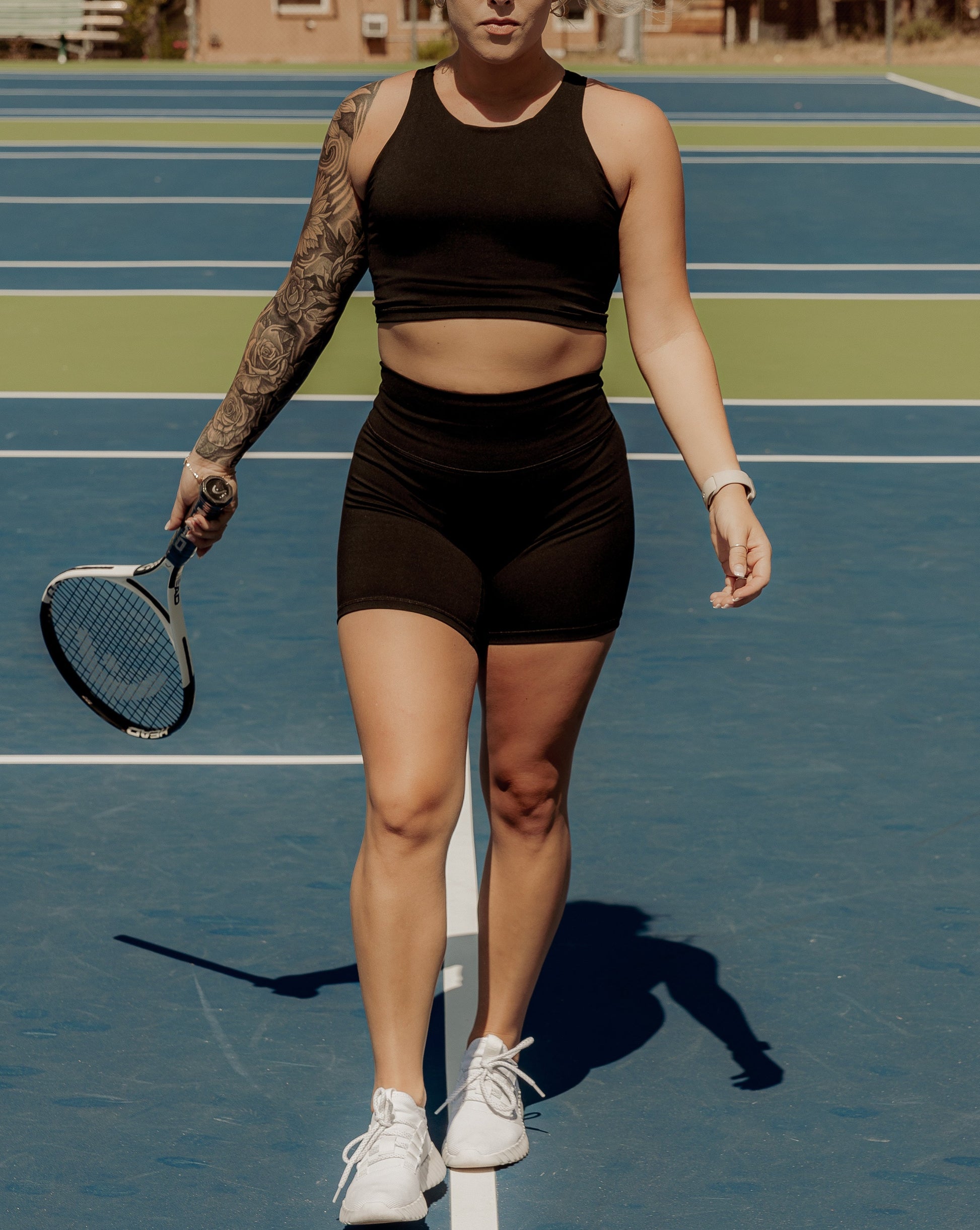 Blonde haired women walking on a tennis court with a tennis racket in her hand.  She is facing the camera and has her hair up.  She is wearing a matching tank top and biker short set in the color black.
