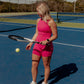 blonde hair women standing in the middle of tennis court holding a tennis racket in her left hand resting it on the ground. she is wearing a matching workout set that includes a long lined tank top and biker shorts in the color hot pink. this photo does not show off her face but her outfit