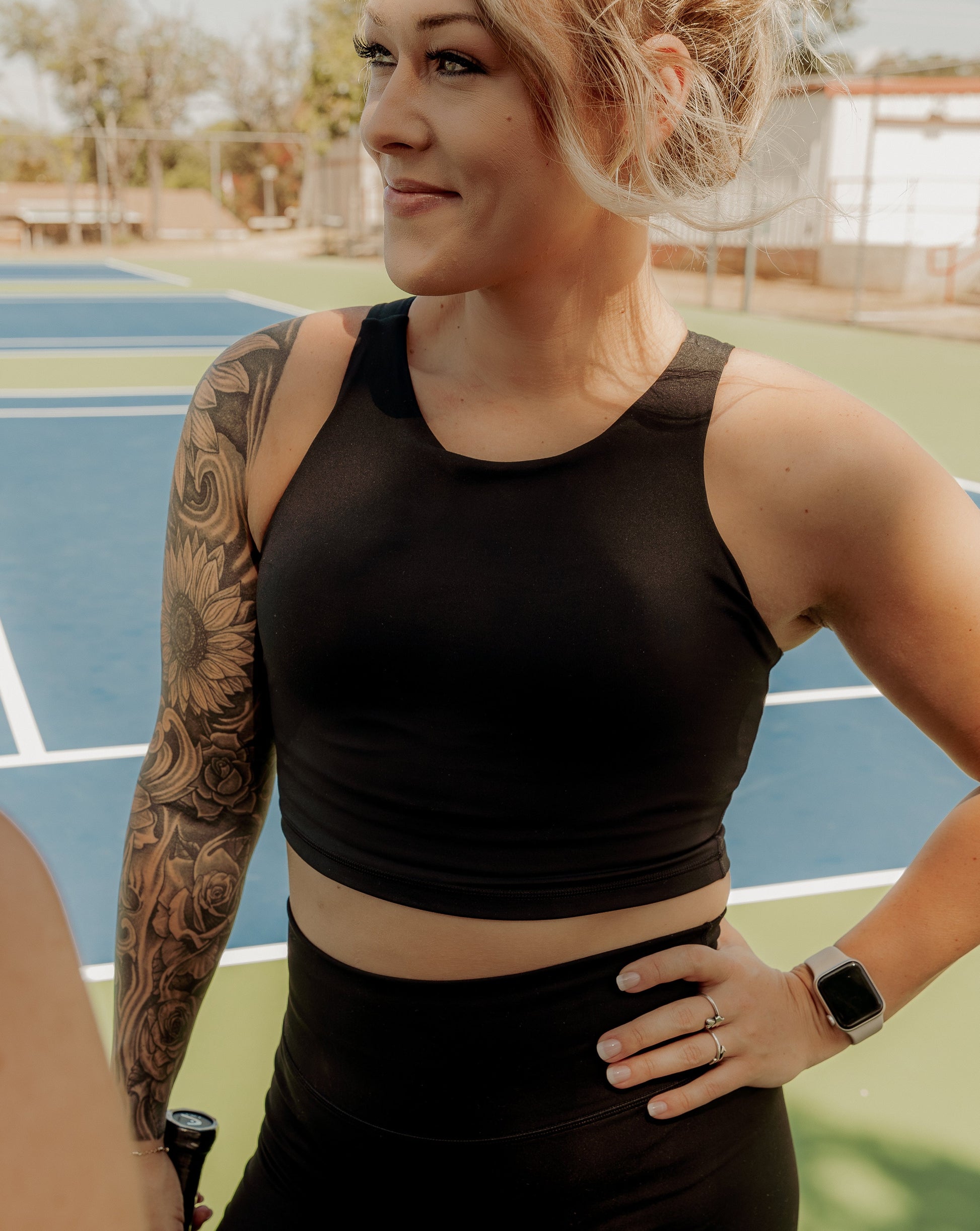 Blonde haired women wearing a matching workout set. Which includes a black tank top and black biker shorts. She is holding a tennis racket and wearing white sneakers standing on a tennis court.