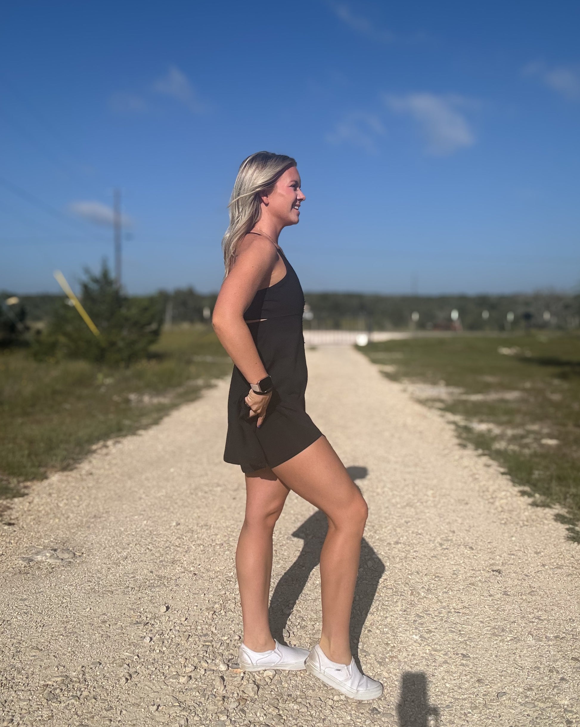 White blonde haired woman wearing a black activewear dress.  She is standing on a dirt road while wearing white vans