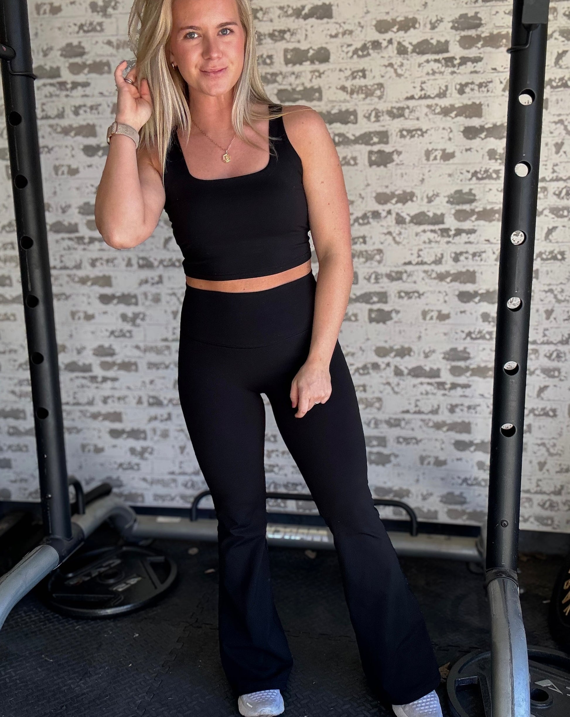white blonde women standing in front of a white washed brick wall.  she is wearing a pair of black flare leggings and a matching long lined tank top with white sneakers.