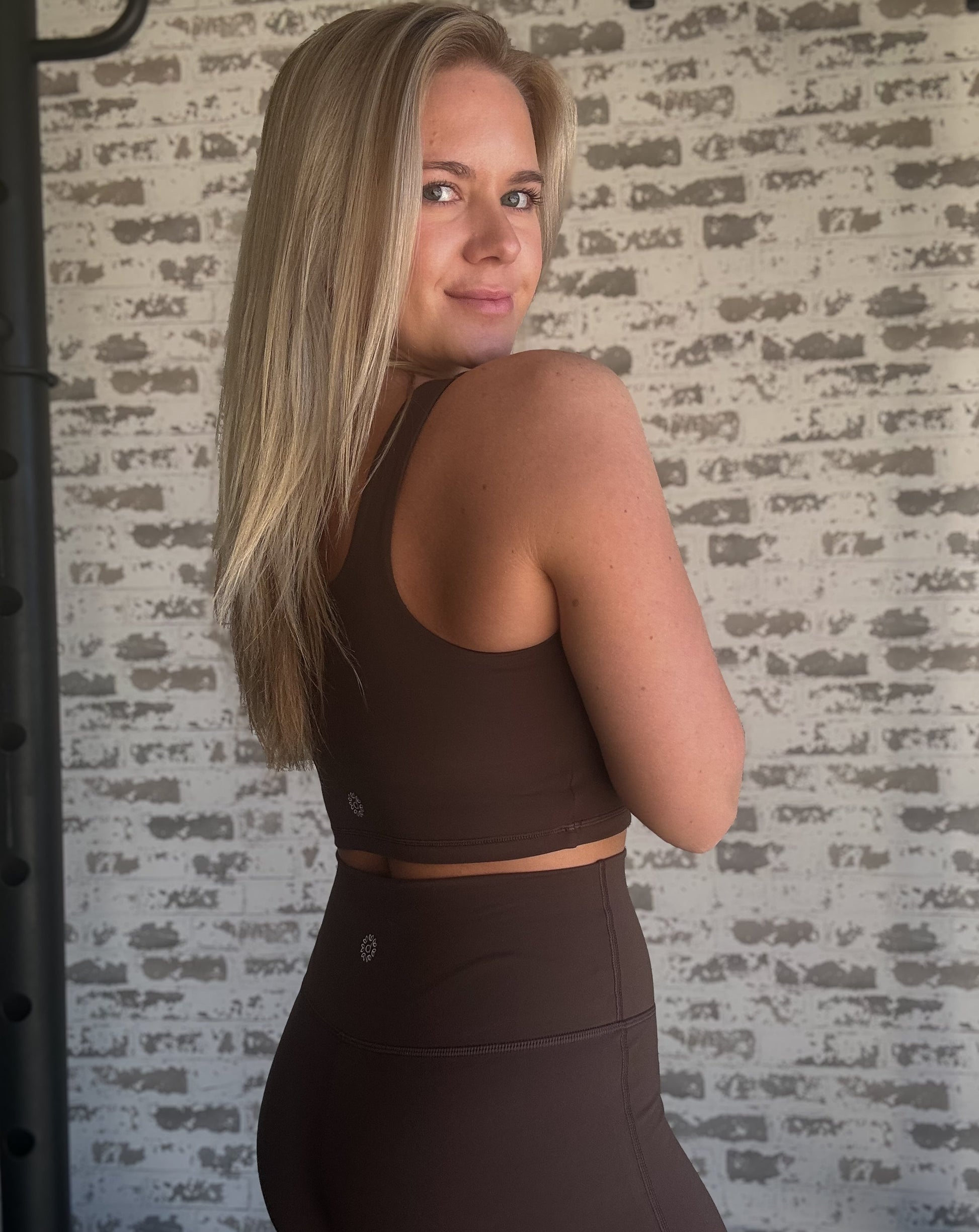 white blonde women standing in front of a white washed brick wall.  she is wearing a pair of brown flare leggings with a matching long lined tank and white sneakers.