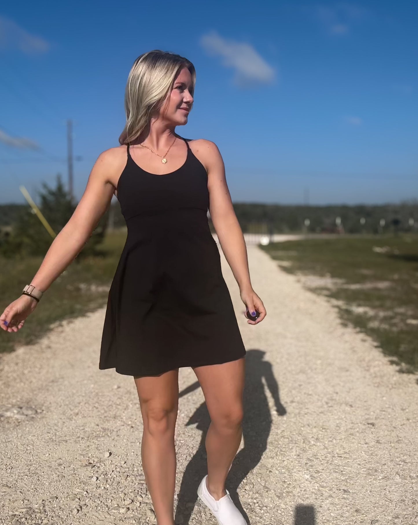 White blonde haired woman wearing a black activewear dress.  She is standing on a dirt road while wearing white vans