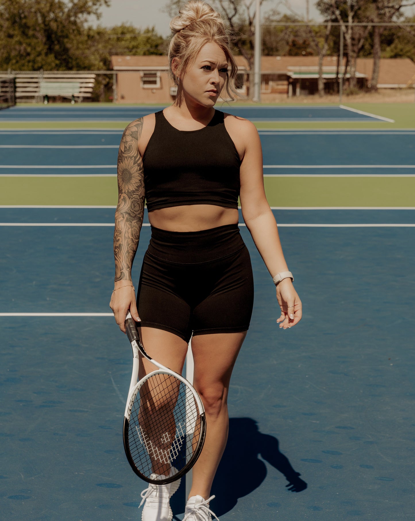 Blonde haired women wearing a matching workout set.  Which includes a black tank top and black biker shorts.  She is holding a tennis racket and wearing white sneakers standing on a tennis court.