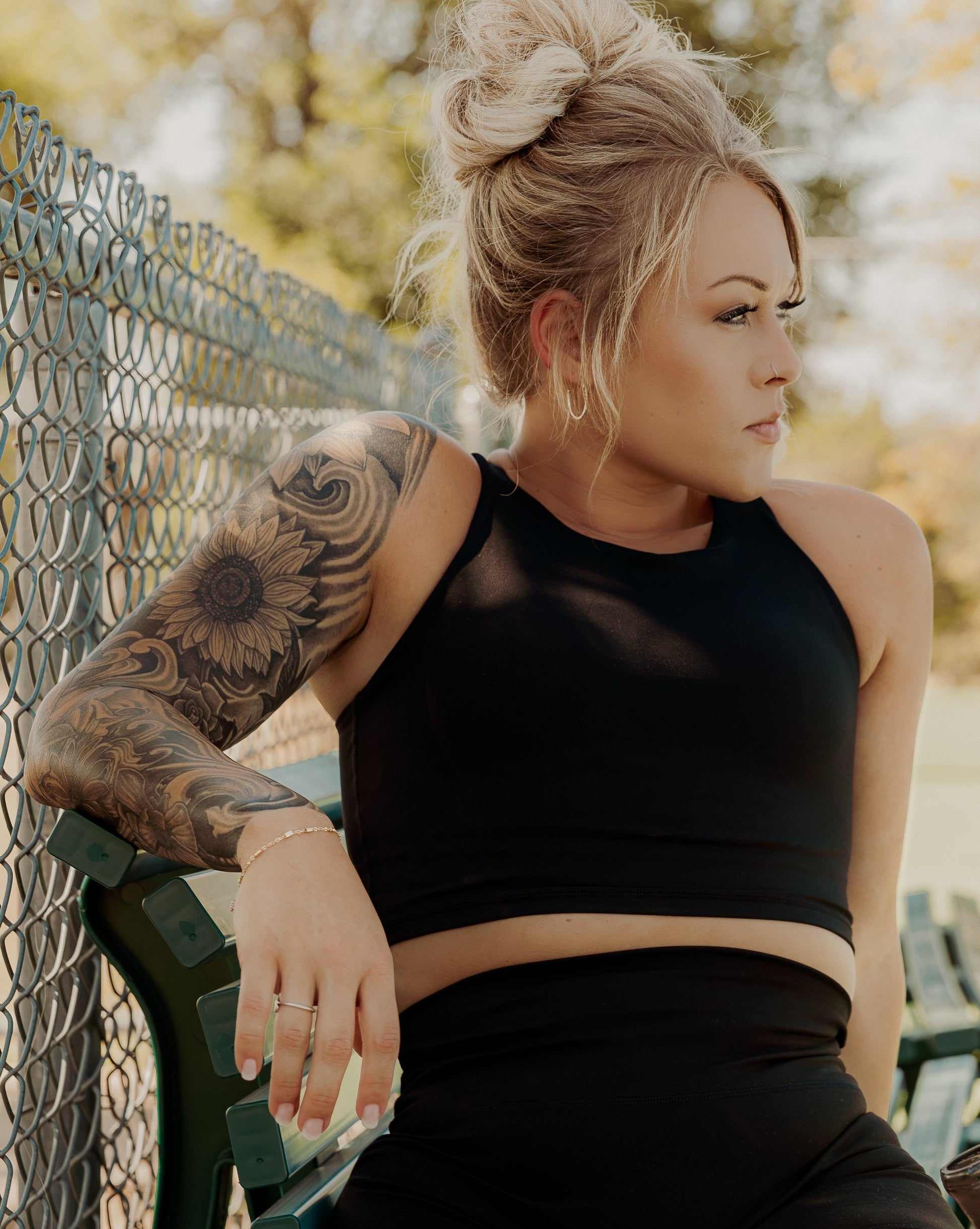 Blonde haired women sitting on a bench at the tennis court.  Her hair is pulled up into a messy bun and she is looking off into the distance.  She is wearing a black tank top with a built in bra and matching black biker shorts.