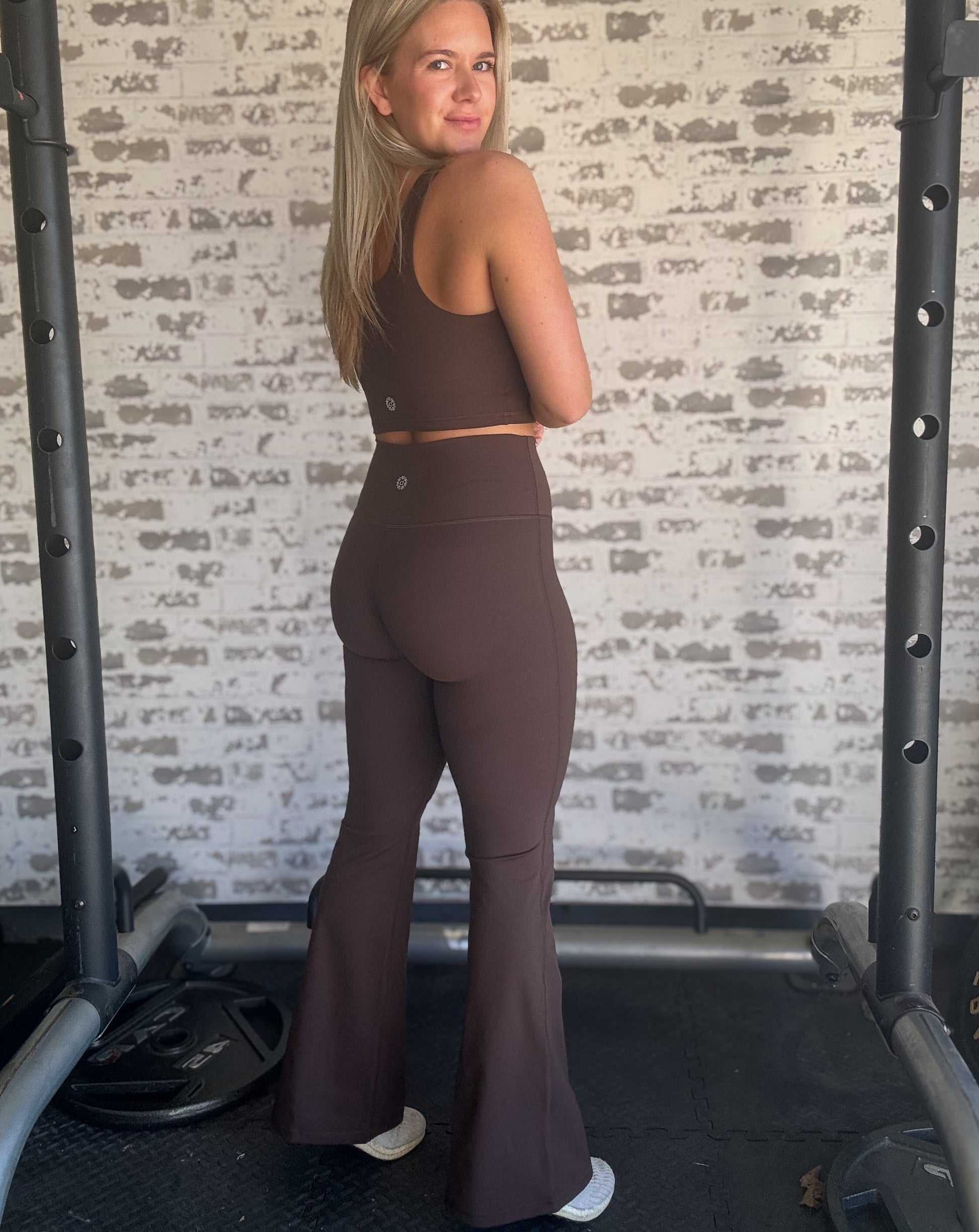 white blonde women standing in front of a white washed brick wall.  she is wearing a pair of brown flare leggings with a matching long lined tank and white sneakers.