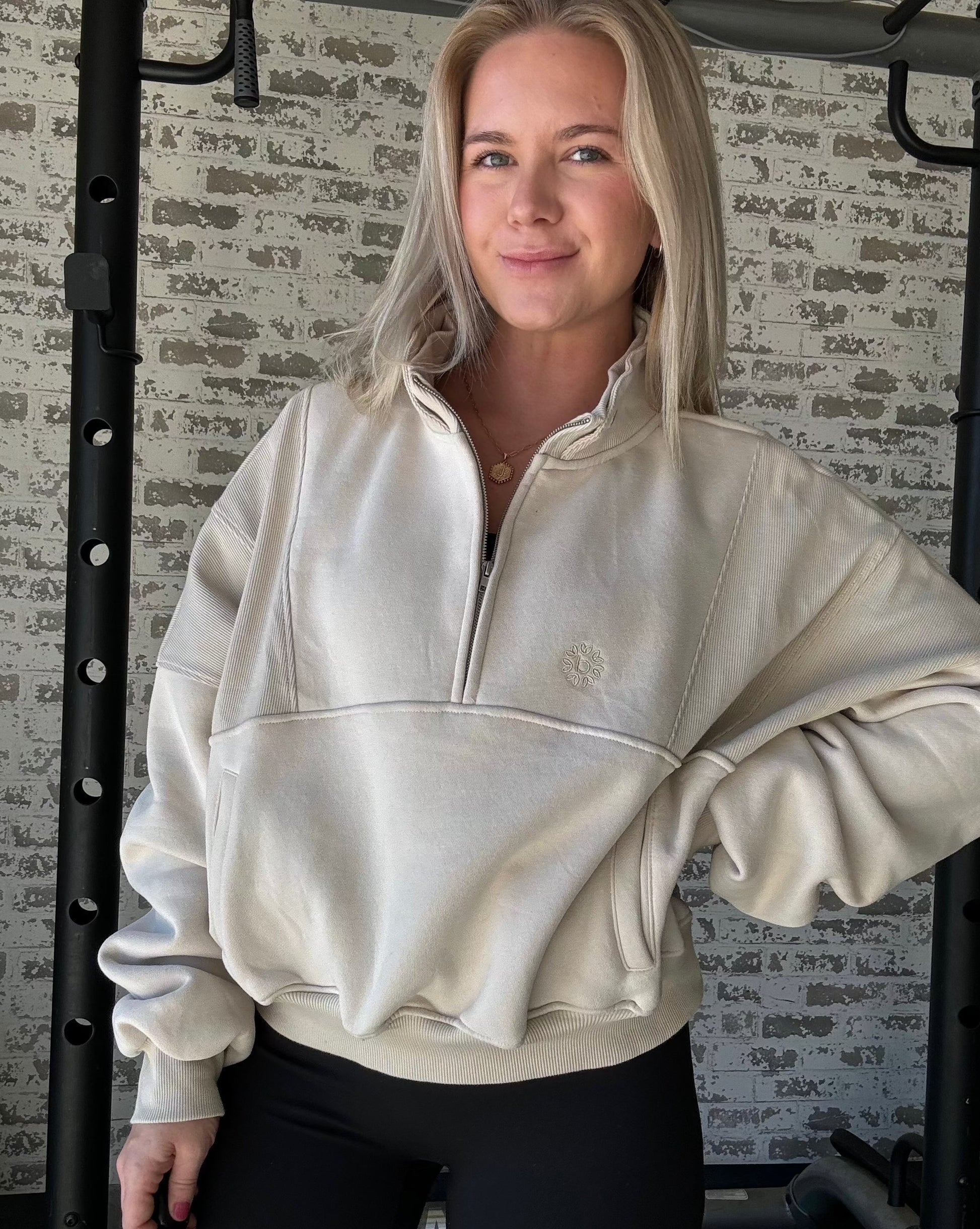 white women wearing an oversized half zip pull over in a tan color.  This women is standing in front of a white washed brick wall.