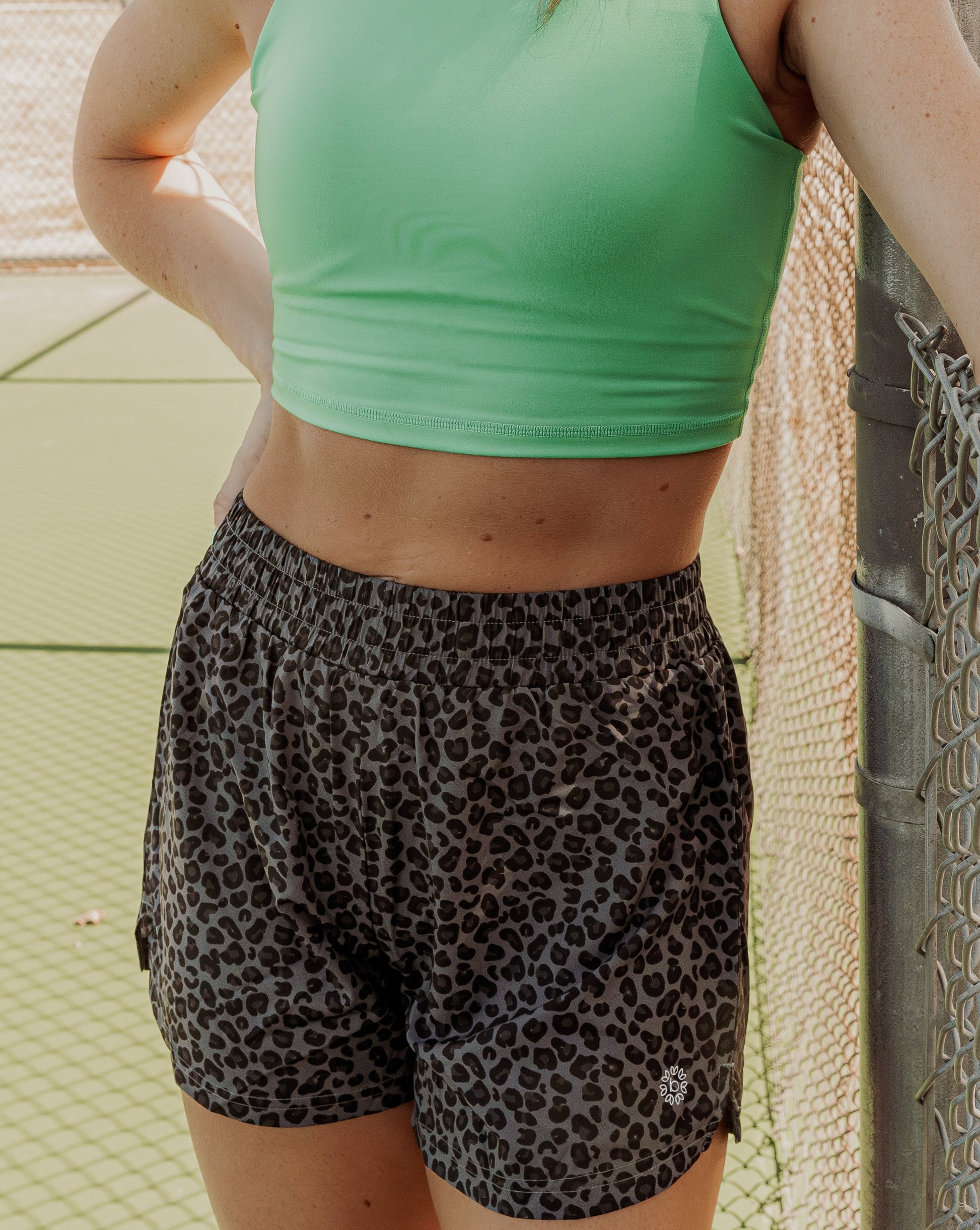 Blonde haired women standing on a tennis court next to the fence.  She is wearing a bright green long lined tank top and high waisted grey leopard running shorts.  This photo does not include her face but the middle half of her body.