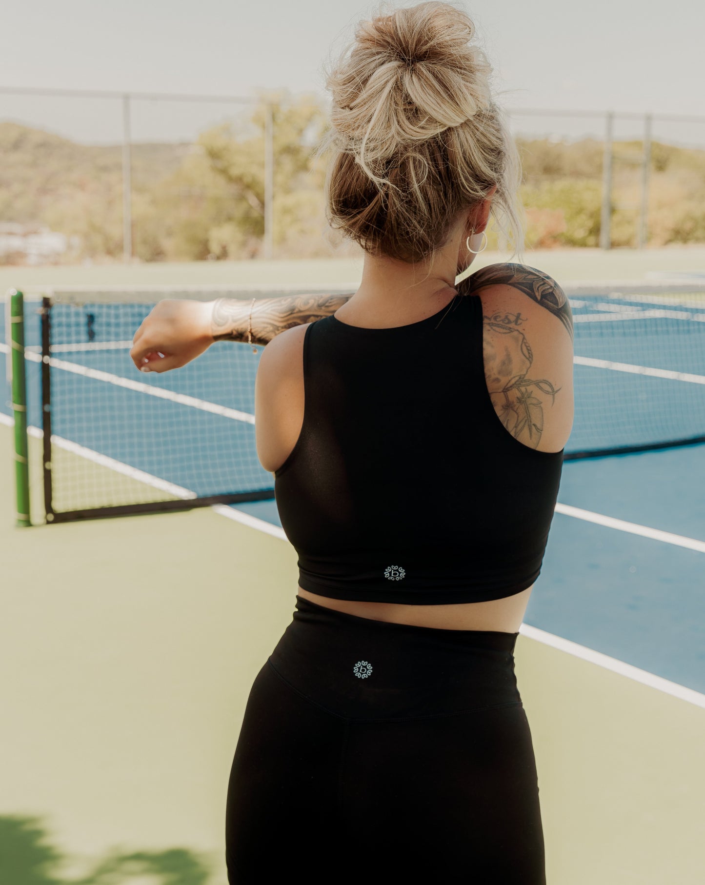 Blonde haired women standing on the middle of a tennis court.  She is facing the opposite direction and is wearing a matching tank top and biker short activewear set.