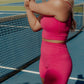 blonde haired women stretching her arms in the middle of a tennis court.  she is wearing a matching hot pink workout set that includes a long lined tank and biker shorts.
