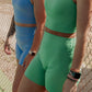 2 blonde haired women standing in front of a chain link fence at a tennis court.  Both are wearing long lined tank tops and matching biker shorts.  One is wearing a bright blue color while the other is wearing a bright green color.