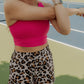 Blonde women wearing a long lined tank top with a built in bra and leopard printed running shorts.  She is streching her arms as she is standing in the middle of a tennis court.