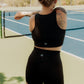 Blonde haired women walking on a tennis court with a tennis racket in her hand.  She is looking away from the camera and has her hair up.  She is wearing a matching tank top and biker short set in the color black.