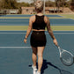 Blonde haired women wearing a matching workout set. Which includes a black tank top and black biker shorts. She is holding a tennis racket and wearing white sneakers standing on a tennis court.