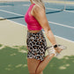 blonde haired women standing in the middle of tennis court wearing a bright pink long lined tank top with a built in bra and leopard printed running shorts.  She is facing to the side and stretching her quads.