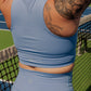 The back side of a women with tatooes on her left arm. She is wearing a matching tank top and biker short set.  She is facing away from the camera and is standing on a tennis court.