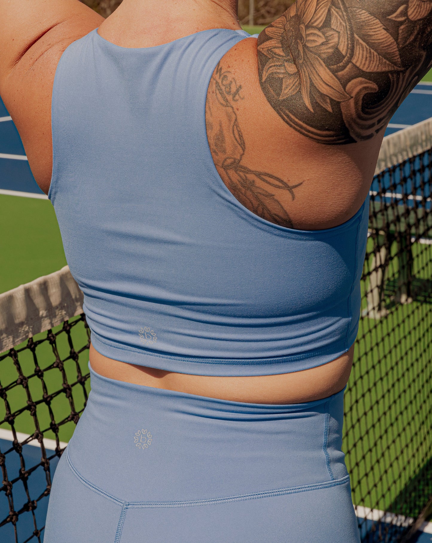 The back side of a women with tatooes on her left arm. She is wearing a matching tank top and biker short set.  She is facing away from the camera and is standing on a tennis court.