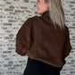 white women wearing an oversized cropped pull over that is a half zip sweater.  She is standing in front of a brick wall with her hands in her pockets of the sweater.