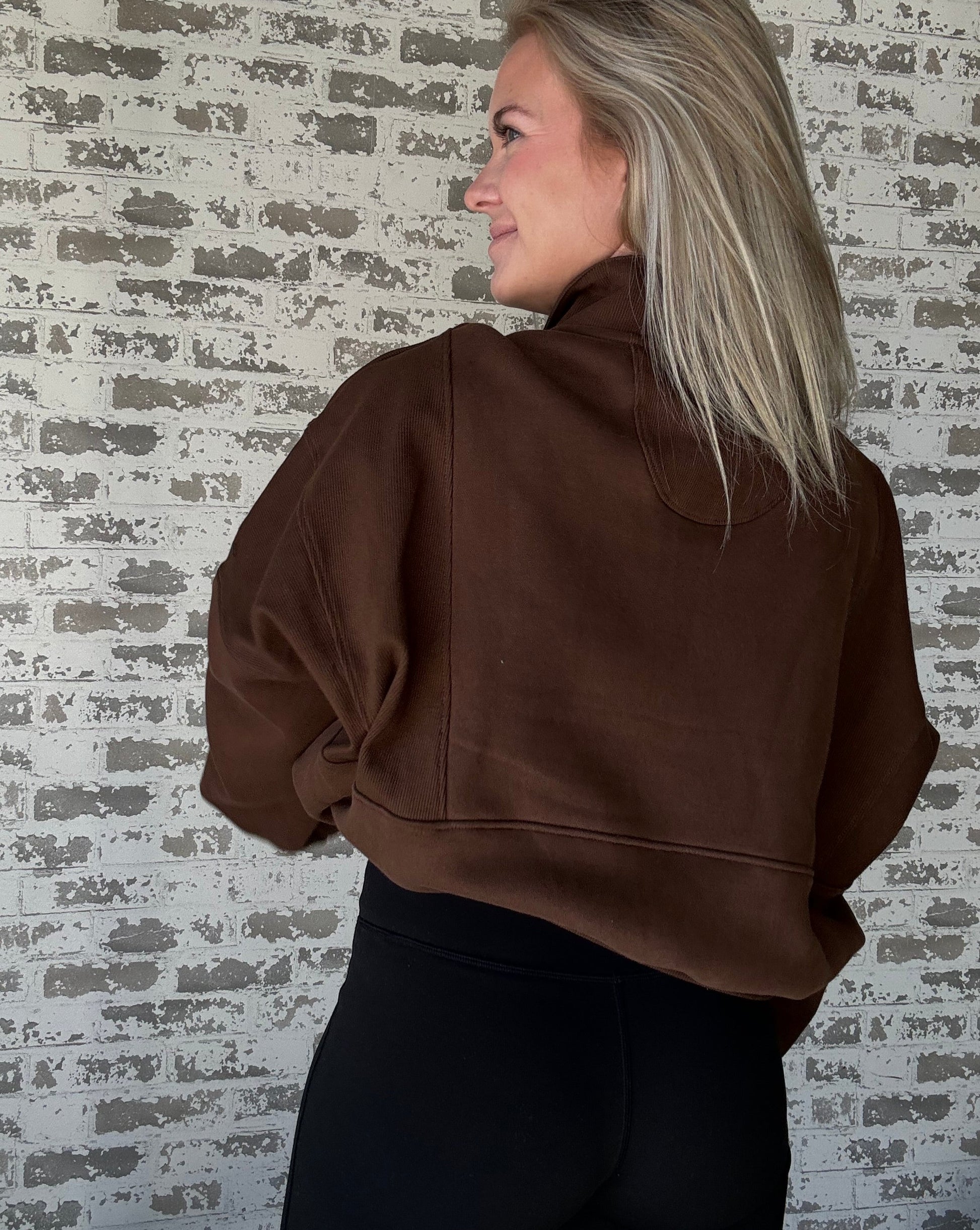 white women wearing an oversized cropped pull over that is a half zip sweater.  She is standing in front of a brick wall with her hands in her pockets of the sweater.