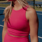Blonde haired women with wavy hair standing on the middle of a tennis court wearing a matching workout set.  This set includes a long lined tank top with a built in bra and matching biker shorts, both in the color bright pink.  This is an up close photo of the tank top from the front.