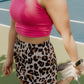 Blonde haired women wearing a bright pink long lined tank top and leopard printed running shorts.  She is stretching her quads while she is standing in the middle of a tennis court.  this photo does not show her face.