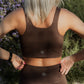 Up close photo of a brown talk top sports bra.  It is being worn by a blonde haired white women who is not facing the camera and has her hands on her hips.  this photo does not show her legs but just her torso