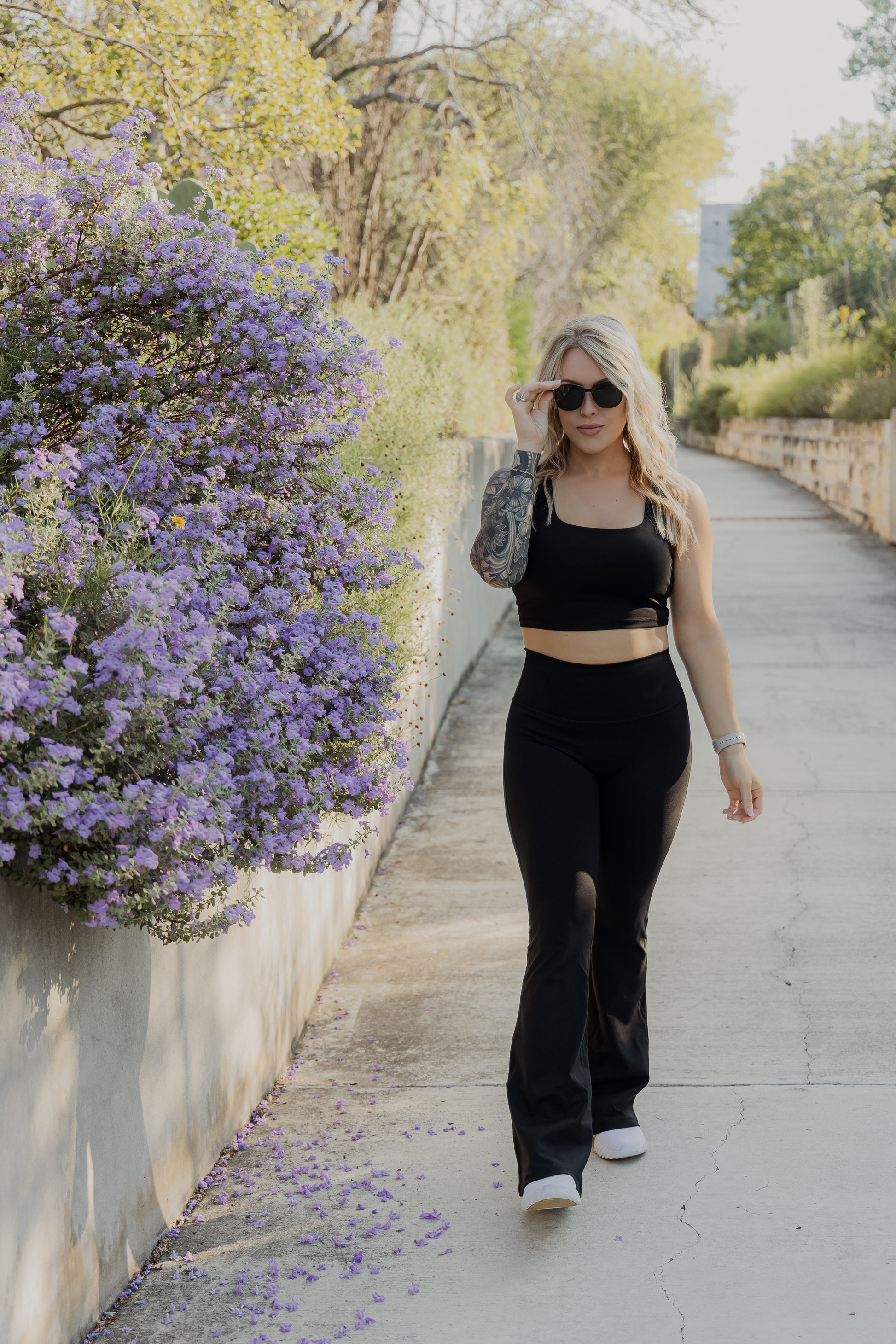 White women with blonde hair wearing a black tank top and flare leggings she is walking down a nature trail area.  She is adjusting the sunglasses on her face.